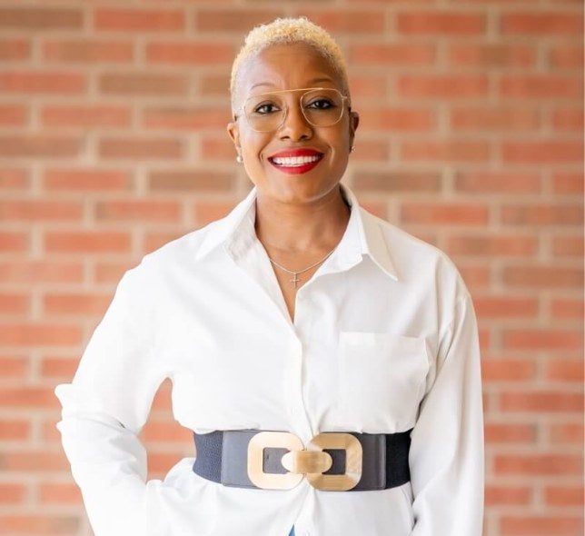 Woman, short hair, glasses, white professional shirt, stands against brick wall.