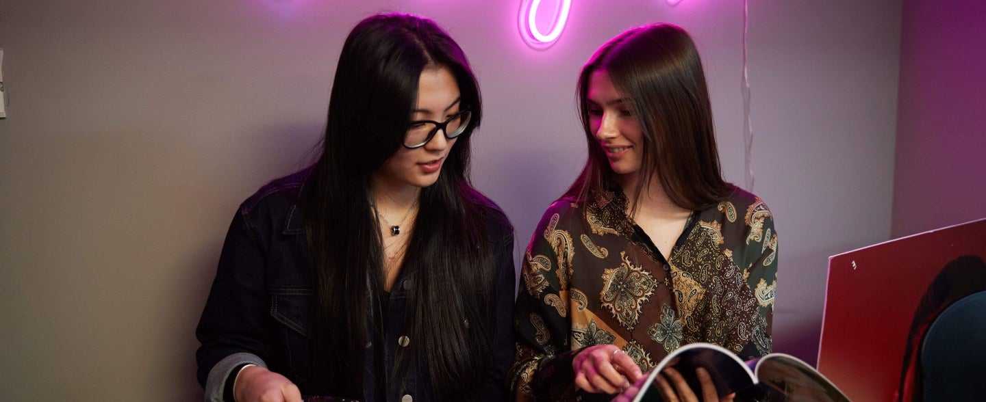 Two LIM students look through the latest issue of The Lexington Line under a neon sign that reads The Lexington Line