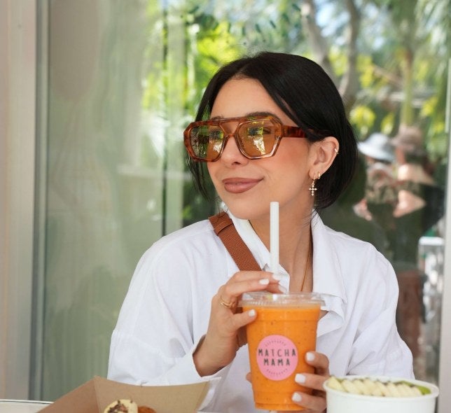 young woman, dark hair, sunglasses, white shirt, drinks an orange Matcha Mama drink