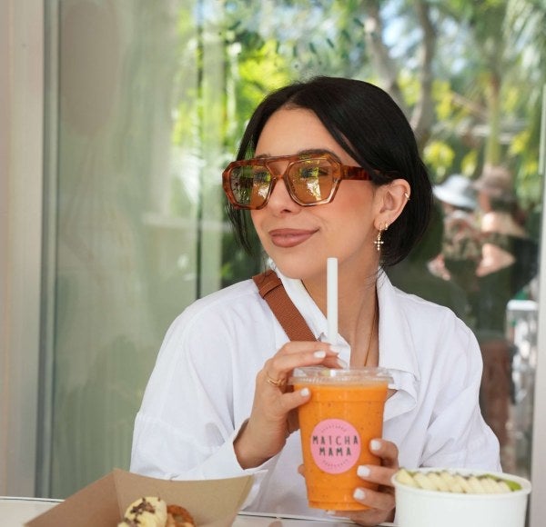 young woman, dark hair, sunglasses, white shirt, drinks an orange Matcha Mama drink