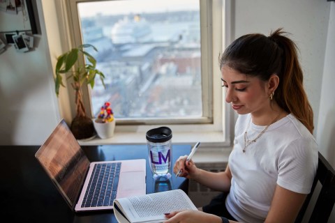 A students sits at a table next to a window with a laptop and book open