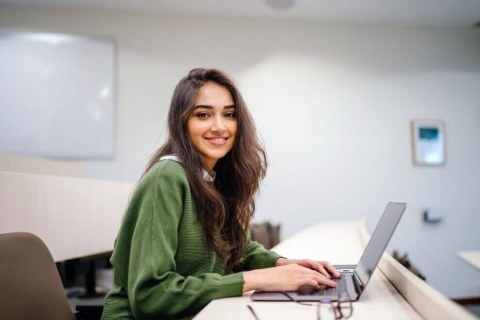 Dark-haired woman smiling over right shoulder, using laptop in a classroom.